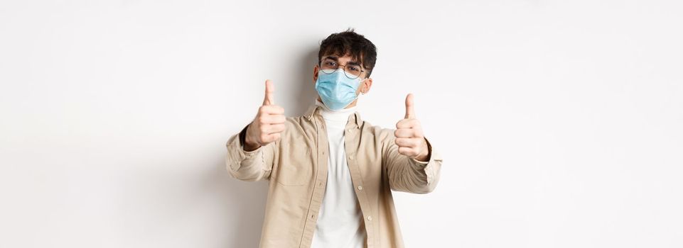 Coronavirus, health and real people concept. Smiling guy in medical mask showing thumbs up, wearing glasses, standing on white background.