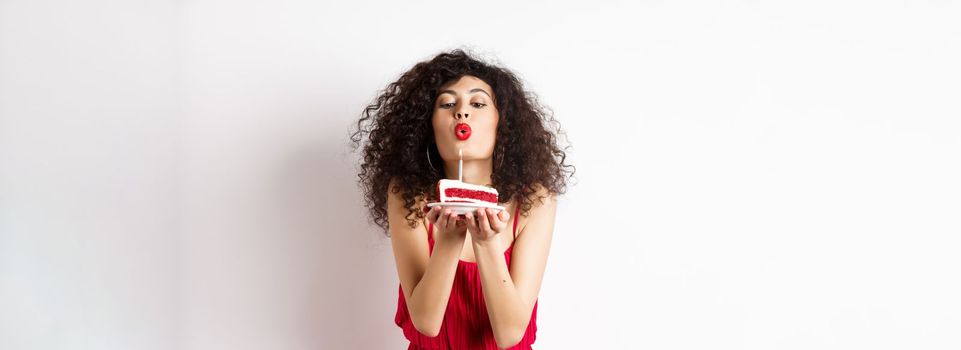 Holidays and celebration concept. Romantic woman in red dress celebrating birthday, blowing out candle on b-day cake and making wish, standing over white background.