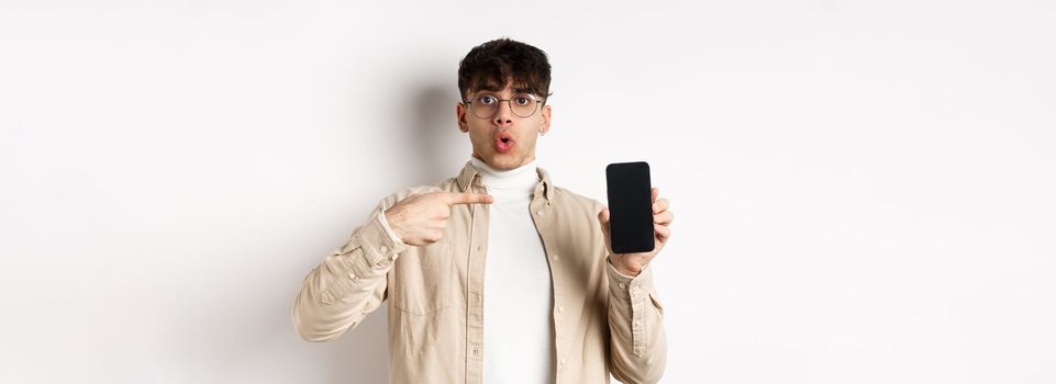 E-commerce concept. Portrait of young man pointing at mobile phone screen, showing advertisement online, standing on white background.
