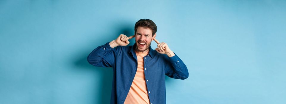 Angry guy shouting with shut ears, block loud music and frowning annoyed, standing on blue background.