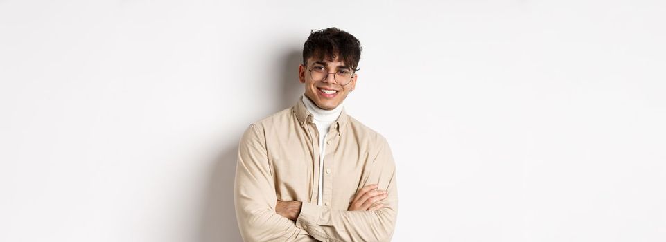 Handsome young man in glasses cross arms on chest, smiling at camera carefree, standing on white background.