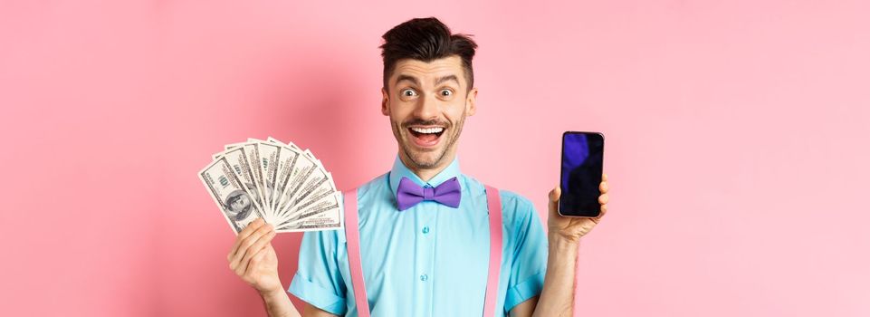 E-commerce and shopping concept. Cheerful guy in bow-tie showing empty smartphone screen and dollars money, smiling amazed at camera, standing on pink background.