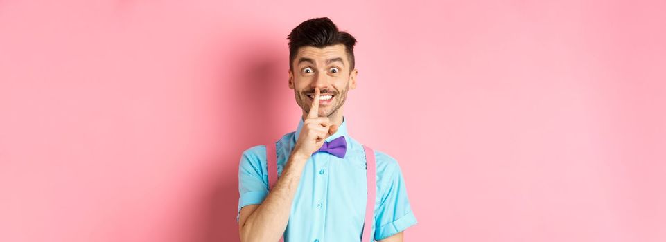 Cheerful young man making surprise, shushing at camera with happy smile, asking to keep voice down, be quiet, standing on pink background.