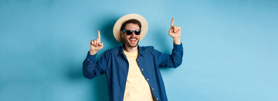 Cheerful caucasian guy in straw hat and sunglasses, dancing and having fun on vacation, standing over blue background. Concept of summer tourism.