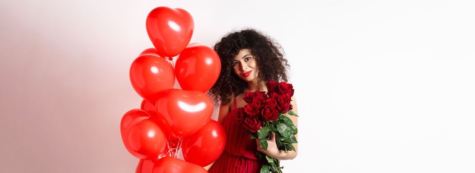 Gorgeous curly haired girlfriend in evening dress, having a date, holding red roses from boyfriend and posing near romantic hearts balloons, white background.