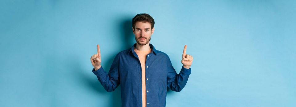 Serious caucasian man with beard pointing fingers up at empty space, showing logo and looking at camera, standing on blue background.