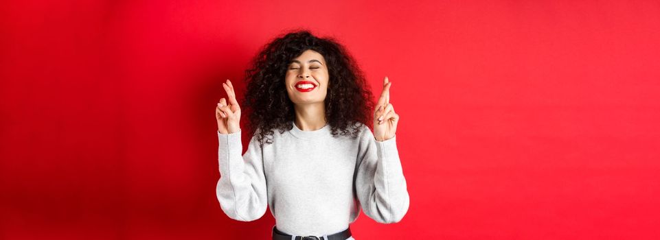 Hopeful and positive woman with red lips and curly hair, cross fingers for good luck and making wish, praying for dream come true, smiling excited, red background.