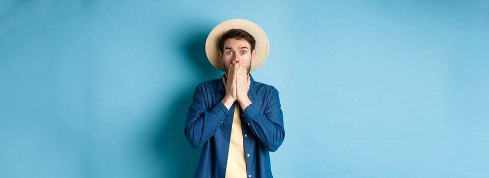 Shocked and alarmed tourist gasping, covering mouth with hands and looking startled at camera, standing on blue background.