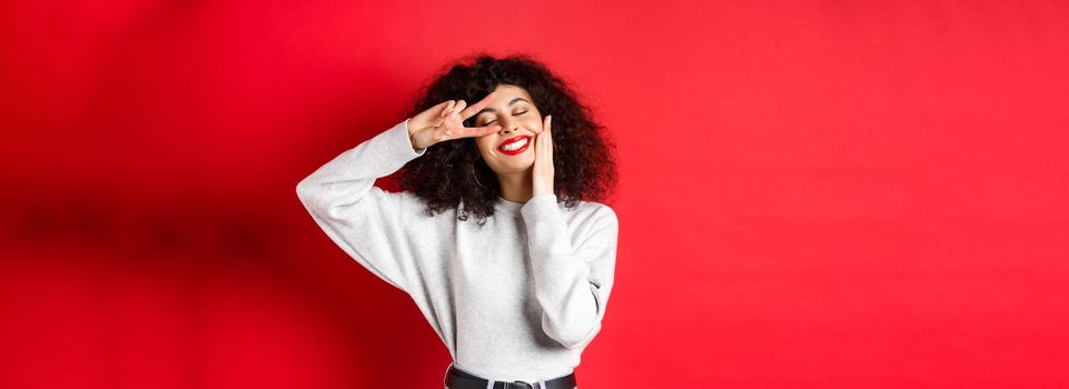 Beauty. Pretty lady with curly hair and red lips, touching face with makeup and showing v-sign on eye, smiling carefree, standing against studio background.