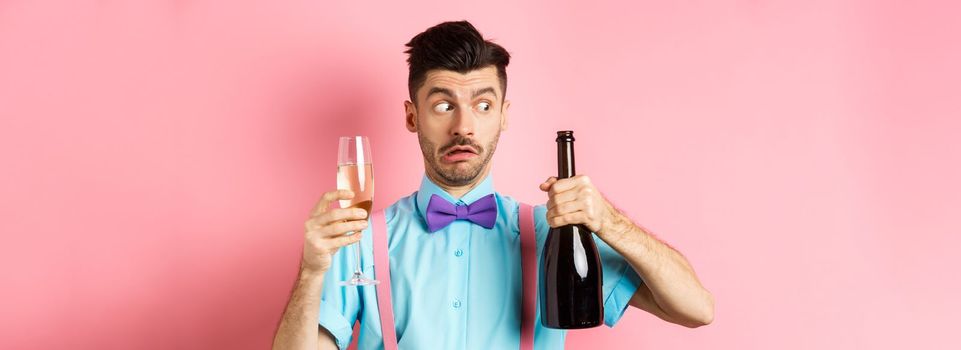 Holidays and celebration concept. Confused drunk guy looking at empty bottle of champagne, holding glass, drinking at party, standing over pink background.