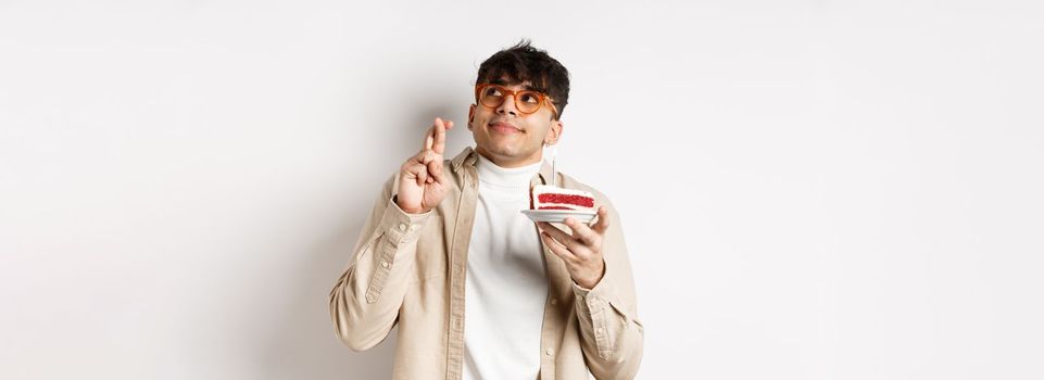 Cute hopeful guy in glasses making birthday wish, holding cake and fingers crossed, looking up and praying, hope dream come true, standing on white background.
