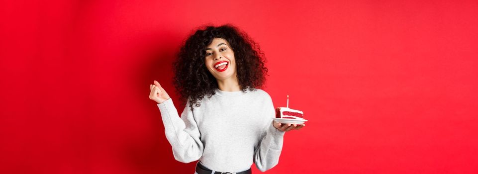 Celebration and holidays concept. Happy beautiful woman dancing and making birthday wish, holding b-day cake and smiling, standing on red background.