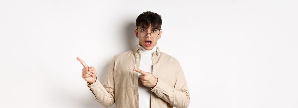 Shocked young man in glasses pointing fingers right at empty space, asking about advertisement, standing on white background.