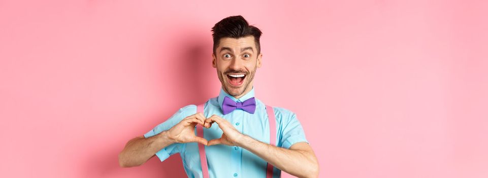 Excited smiling man showing pounding heart and looking with love, standing over romantic pink background. Valentines day concept.