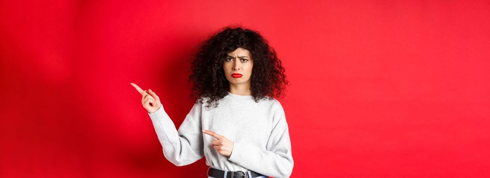 Sad and gloomy brunette woman with curly hair, frowning and looking disappointed, pointing fingers right at logo, standing on red background.