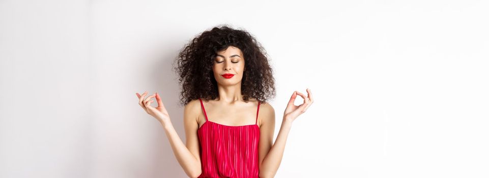 Calm and relaxed smiling woman in red dress, close eyes and meditating, practice yoga in zen pose, standing on white background.