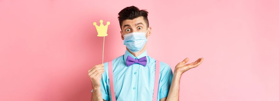 Covid, pandemic and quarantine concept. Clueless and confused young man in medical mask shrugging, showing little party crown, know nothing, standing on pink background.