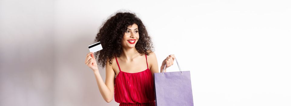 Beautiful woman with curly hair, red dress, showing shopping bag and plastic credit card, white background.