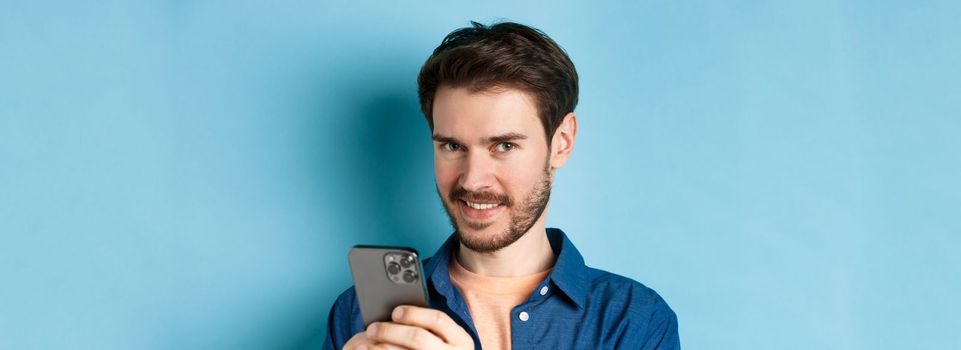 Close-up of attractive caucasian man holding mobile phone and smiling at camera, standing in casual outfit on blue background.