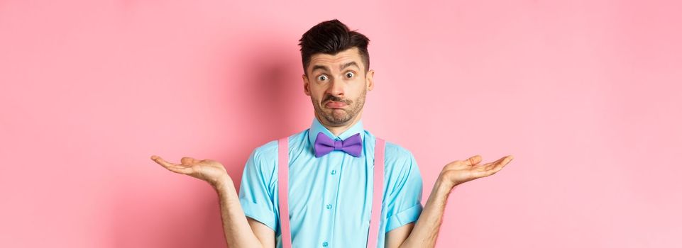 Image of confused guy in bow-tie and suspenders know nothing, shrugging shoulders and looking clueless, standing over pink background.