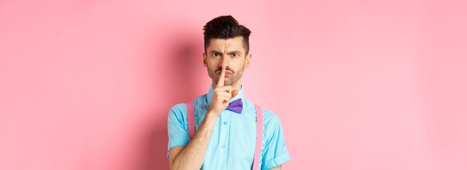 Angry and grumpy man in funny bow-tie shushing at camera, frowning and demand silence, tell to be quiet, scolding someone loud, standing on pink background.