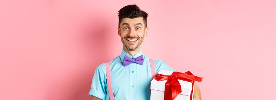 Holidays and celebration concept. Cheerful guy in bow-tie and suspenders bring gift box to party, holding present and smiling, standing over pink background.