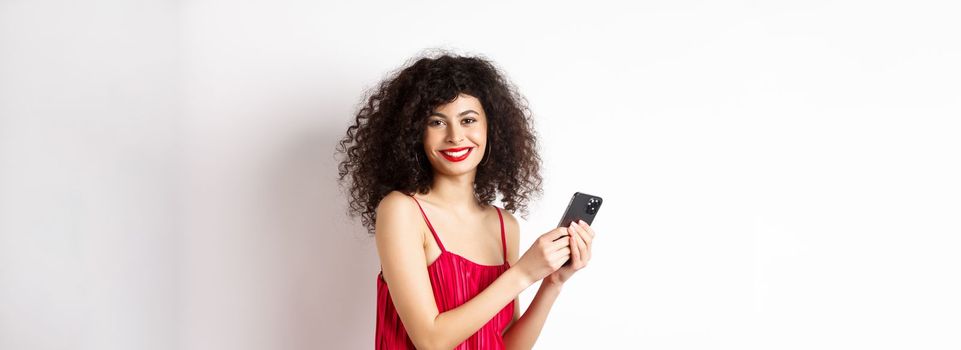 Happy elegant woman in red dress writing message, using smartphone and smiling at camera, standing against white background.