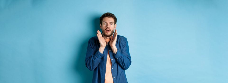 Shocked and scared young man standing in stupor, raising hands up and gasping speechless, standing on blue background.