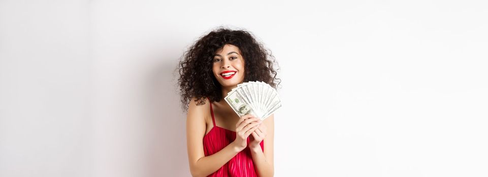 Excited woman in red dress winning money, showing dollar bills and smiling happy, standing on white background.