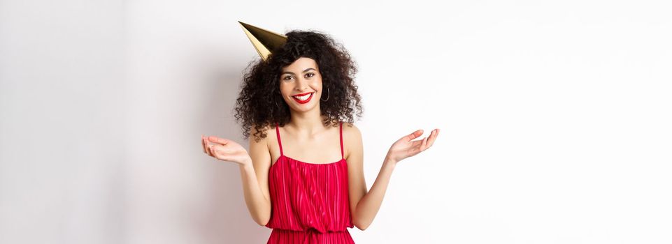 Beautiful caucasian woman in red dress, celebrating holiday, wearing party hat and smiling, standing on white background. Copy space