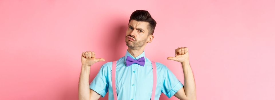 Serious and funny young man trying look cool, pointing at himself to self-promote, being a professional, standing over pink background.