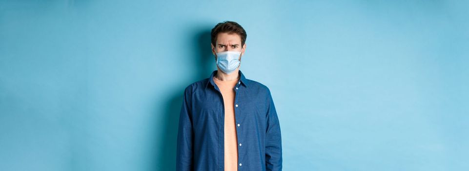 Covid-19 and healthcare concept. Disappointed guy in medical mask frowning, looking confused, standing on blue background.