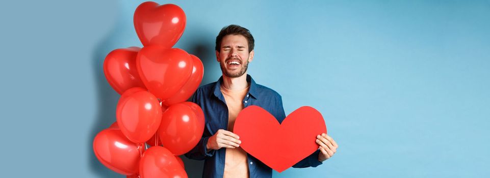 Heartbroken man crying of breakup of valentines day, holding red heart cutout and standing near romantic balloons over blue background.