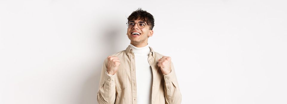 Lucky young man say yes with relieved face, smiling and making fist pump, triumphing of success, winning prize, standing on white background after achieving goal.