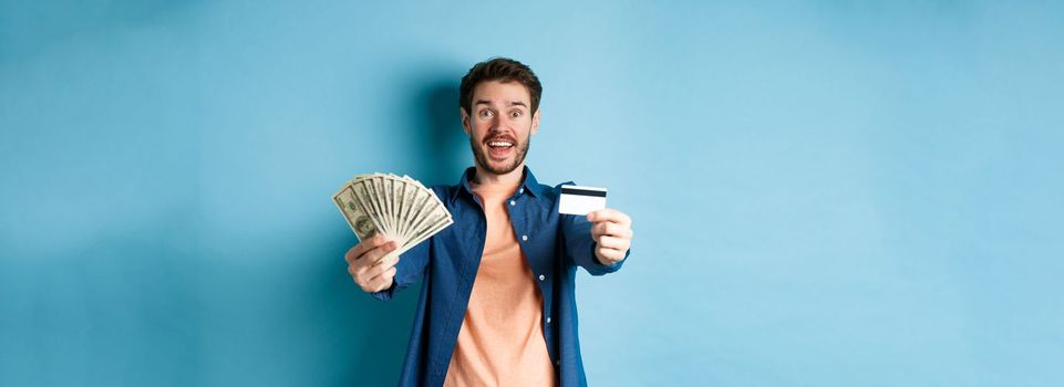 Cheerful caucasian guy extending hands with dollars and plastic credit card, smiling happy at camera, standing on blue background.