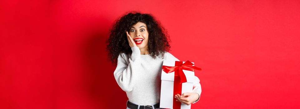 Surprised pretty girl receive Valentines day romantic gift, holding present box and looking with disbelief and happiness at camera, standing on red background.