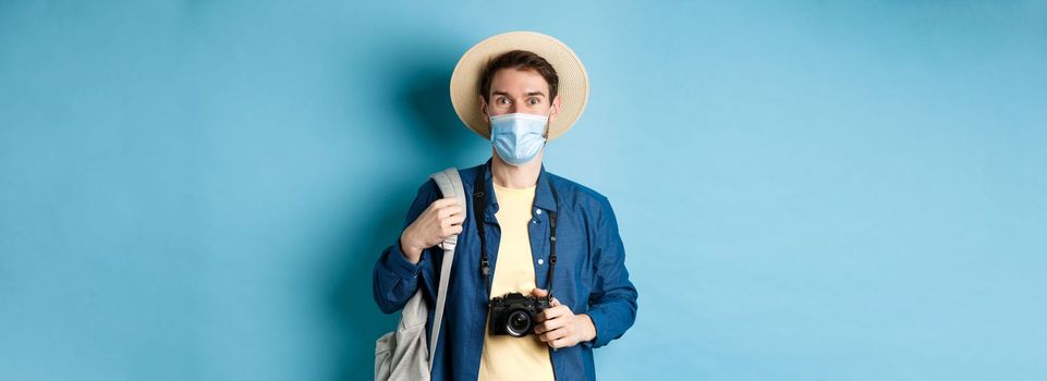 Covid-19 and travelling concept. Young guy tourist in medical mask and summer hat travel abroad during coronavirus pandemic, taking pictures on vacation, blue background.