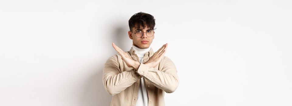 Image of serious young guy in glasses say no, showing cross gesture, make stop sign to disagree or prohibit something, standing on white background.