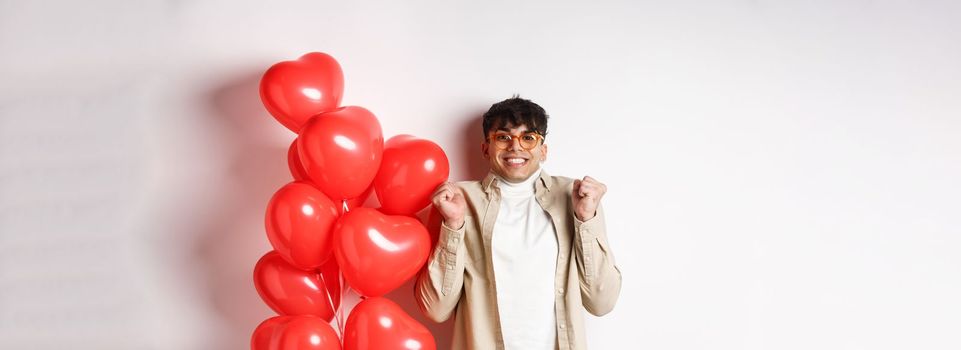 Valentines day. Excited smiling man eager to go on date, celebrating with lover, standing near red hears balloons, white background.