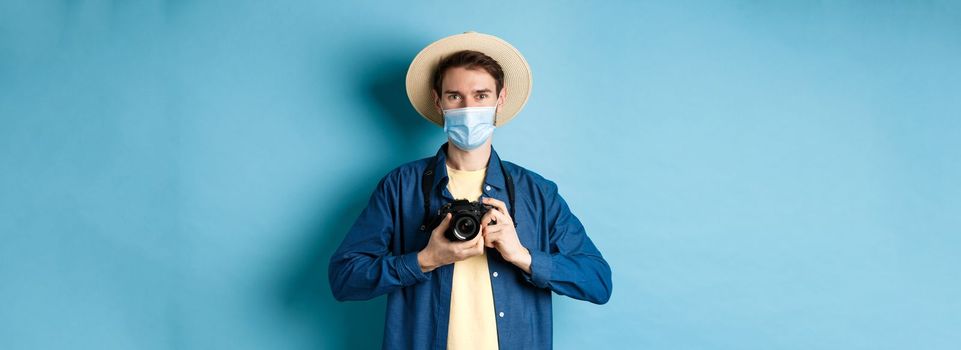 Covid-19, pandemic and travel concept. Happy guy on vacation taking pictures with camera, wearing medical mask and summer hat, blue background.