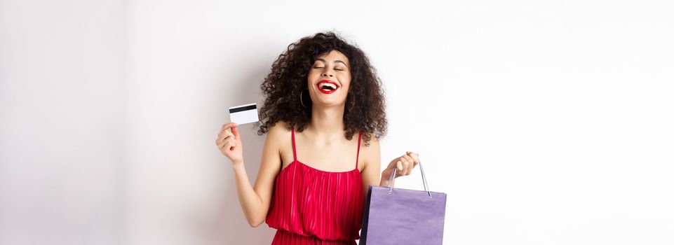 Happy woman in red dress, laughing and shopping, holding plastic credit card and store bag, standing on white background.