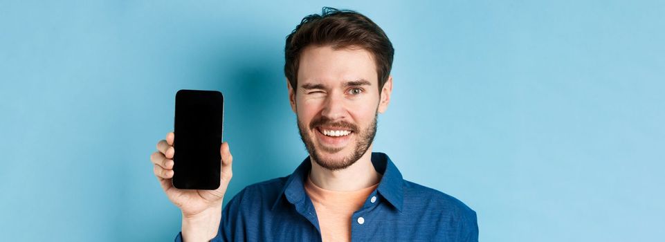 Close-up of handsome young man smiling, winking and showing empty mobile phone screen, standing in casual clothes on blue background.