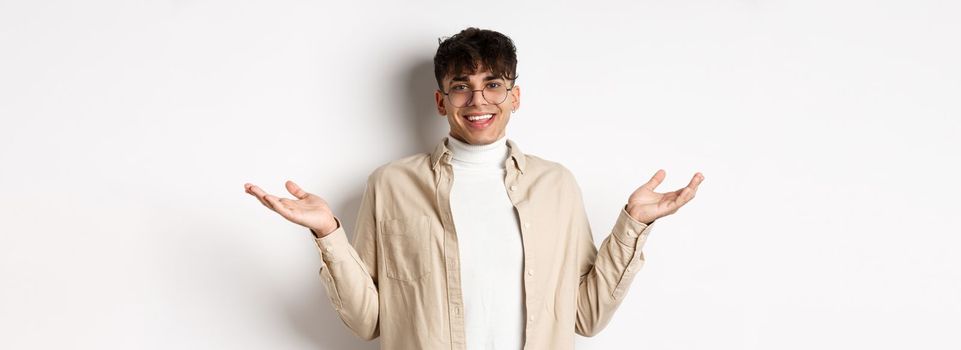 Portrait of happy and pleased young man spread hands sideways and smiling friendly, looking satisfied, standing in glasses on white background.