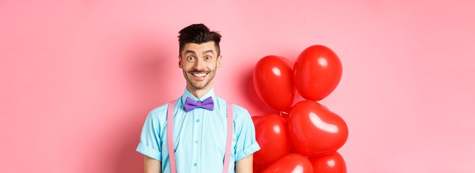 Valentines day concept. Image of handsome young man looking excited and surprised, smiling while standing on romantic pink background near heart balloons.