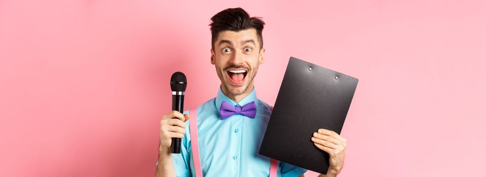 Holidays concept. Happy young man entertainer smiling at camera, holding clipboard and microphone, making speech on party event, standing over pink background.