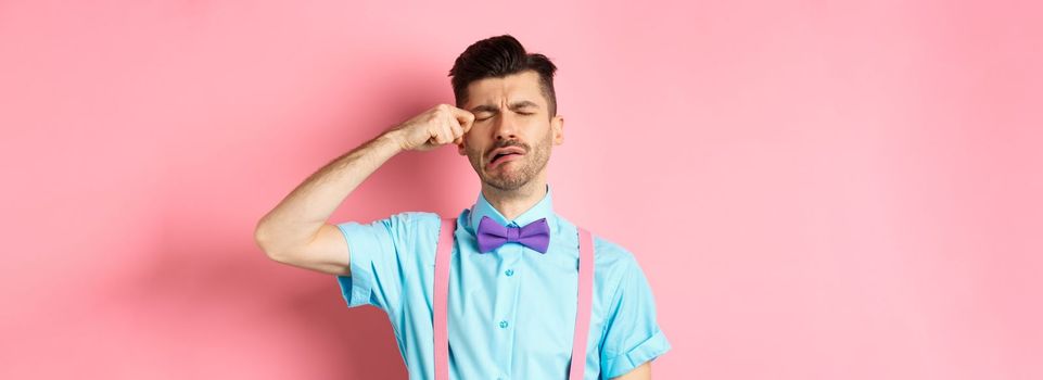 Image of heartbroken guy crying and wiping tear off face, sobbing and feeling sad or lonely, standing upset on pink background.