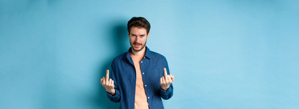 Ignorant and unbothered man showing middle-fingers and looking reluctant at camera, give zero fucks, standing on blue background.