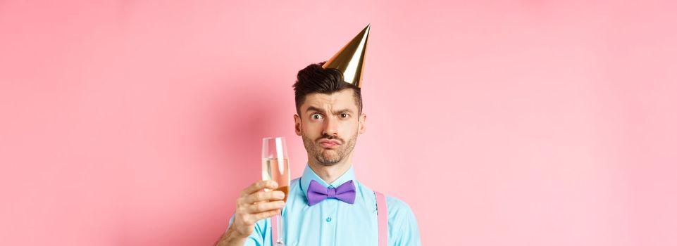 Holidays and celebration concept. Troubled young man in party hat, frowning with doubtful face, raising glass of champagne perplexed, standing on pink background.