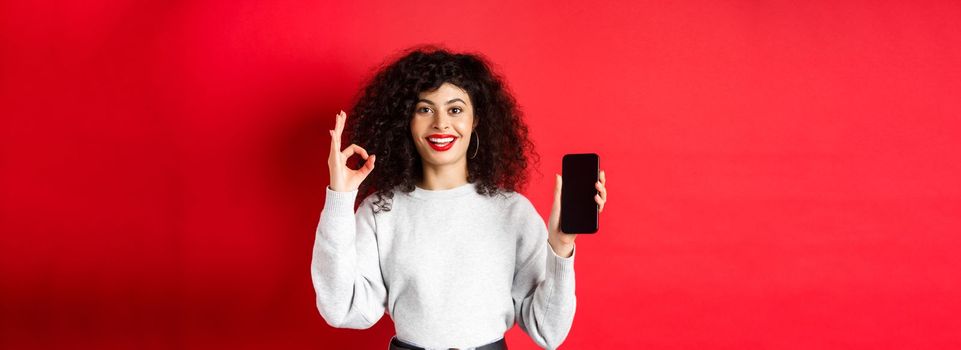 Cheerful european woman with curly hair, showing empty mobile phone screen and okay gesture, smiling satisfied, praise good app or promotion, red background.