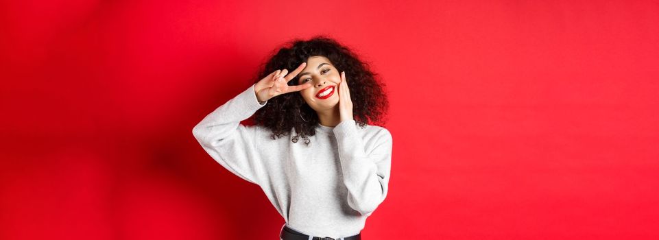 Beauty and makeup. Happy young woman with curly hair, touching face and showing v-sign with cute smile, standing against red background.